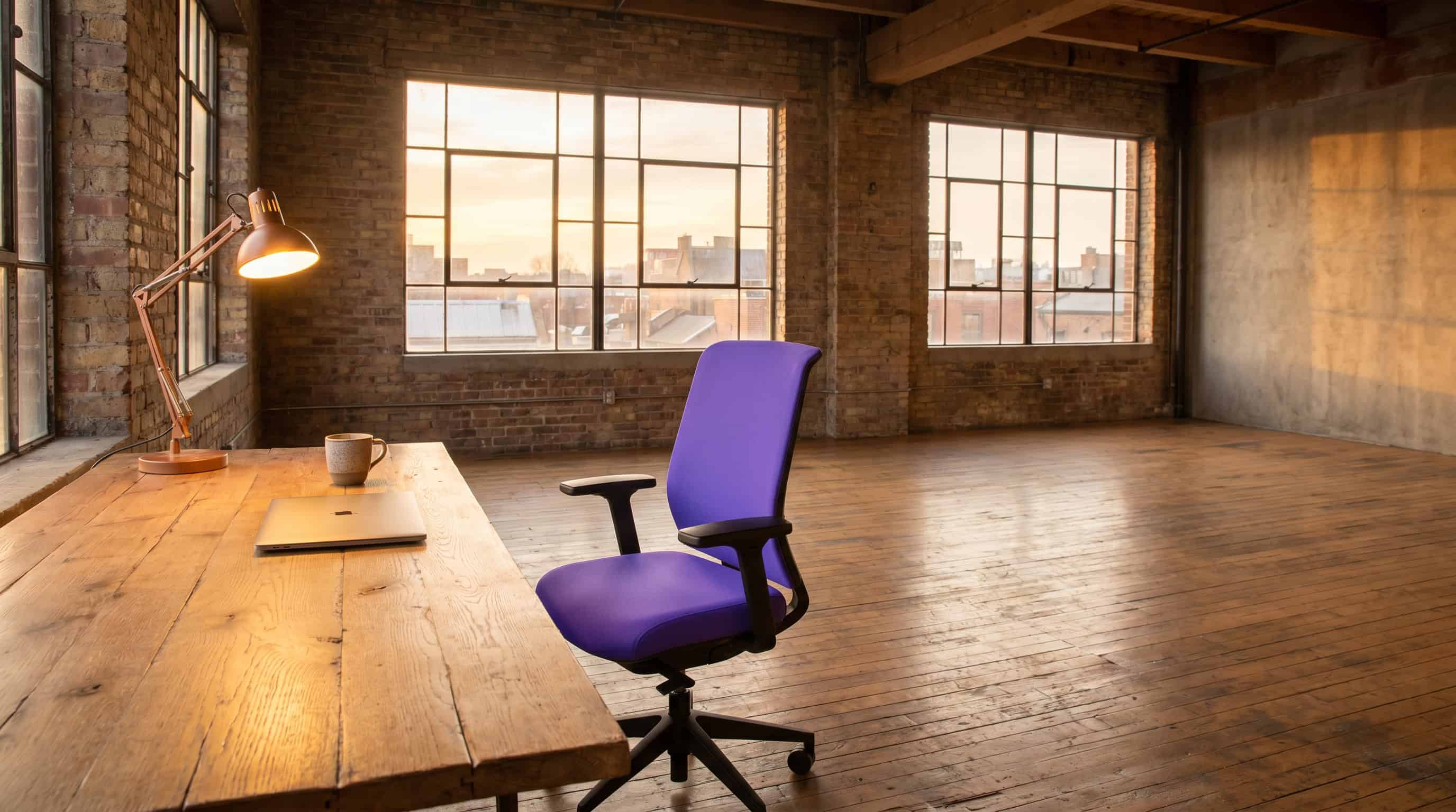 Empty brick-loft studio at sunset with a desk lamp, ergonomic chair, laptop, and coffee cup on an oak desk.